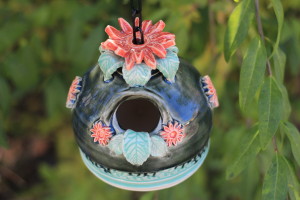 This is a two toned green/blue small bird house with a fancy orange mum flower on the top and smaller flowers along the sides. The green leaves from the opening serves as a perch for our little feather mom to feed her babies from! 